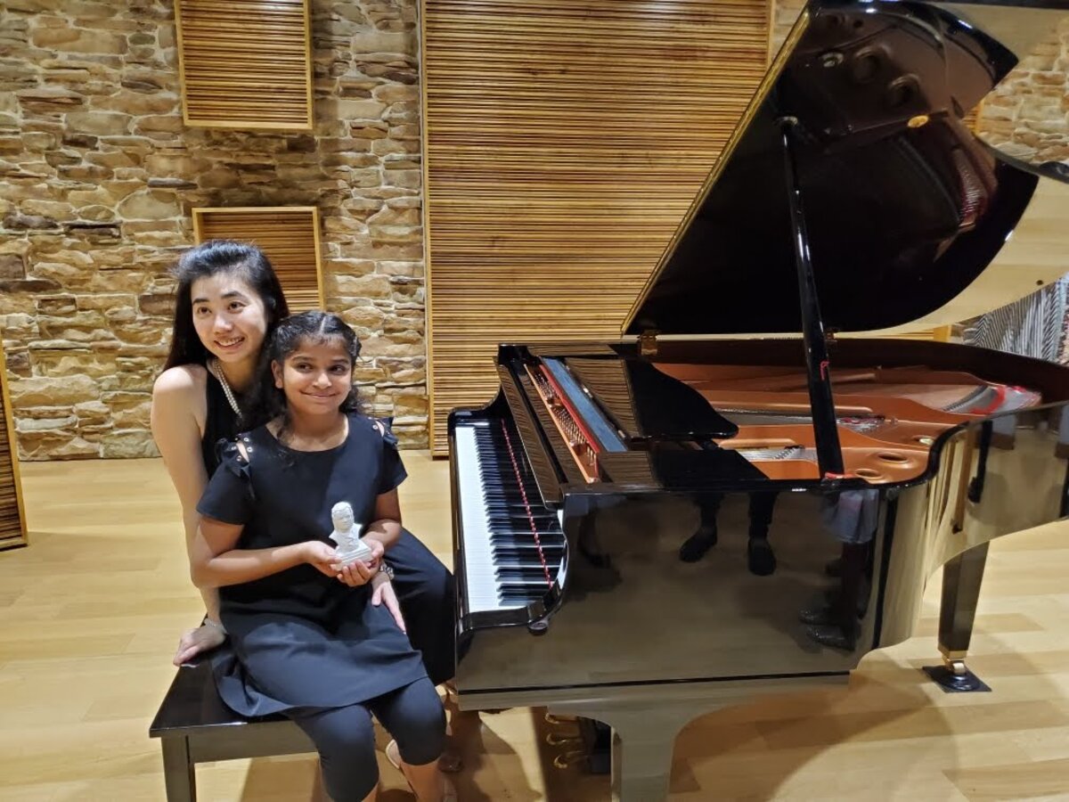Aahana with her piano instructor at a grand piano, holding a recital award